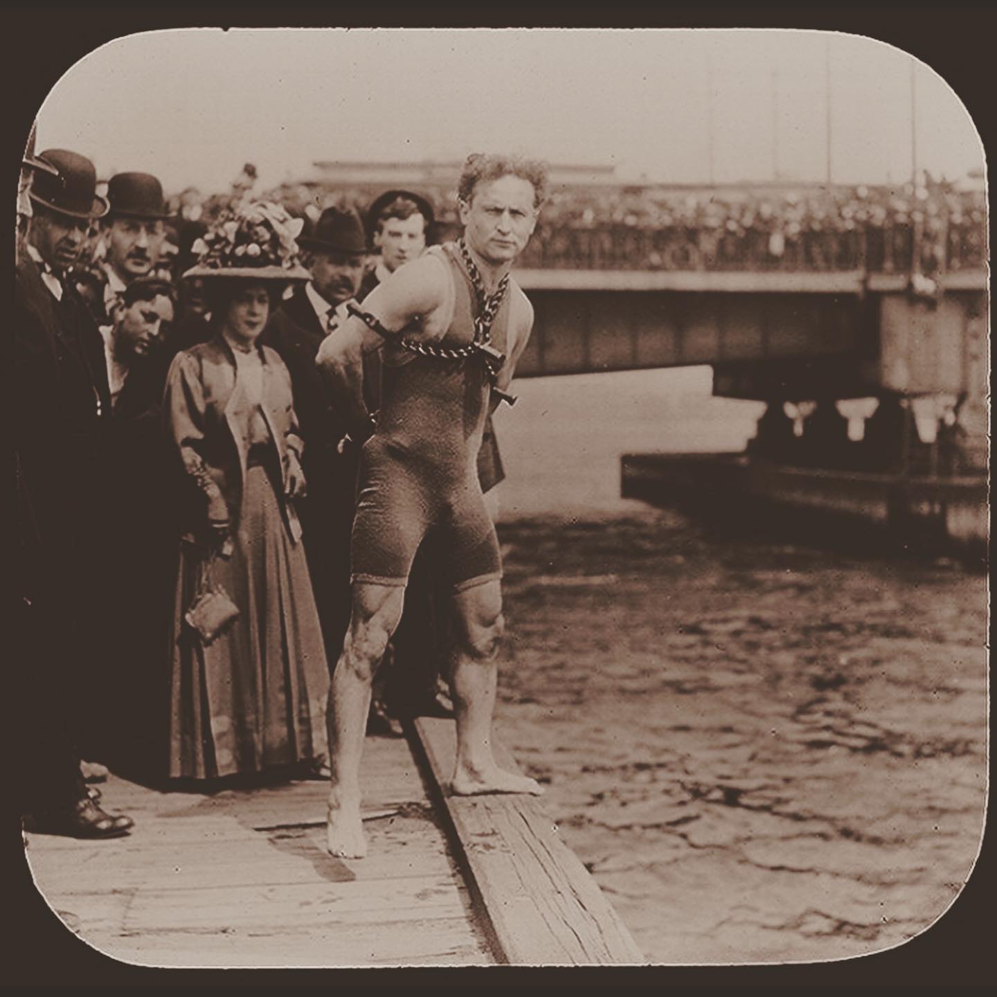 Harry Houdini in chains, poised above the river before an escape — a crowd watching from the bridge