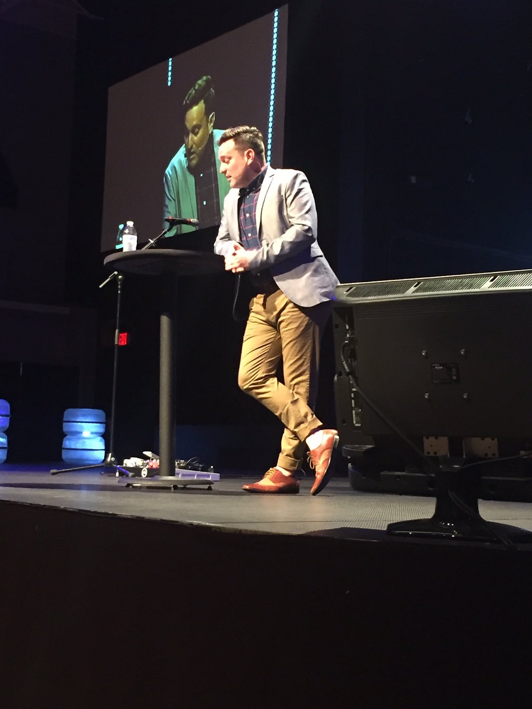 Stephen Poore speaking on stage under stage lights, his image projected on the screen behind him